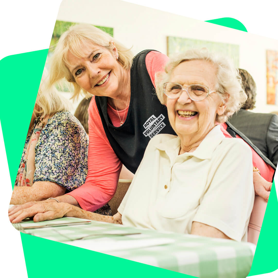 A volunteer leaning over a older lady at a lunch club 
