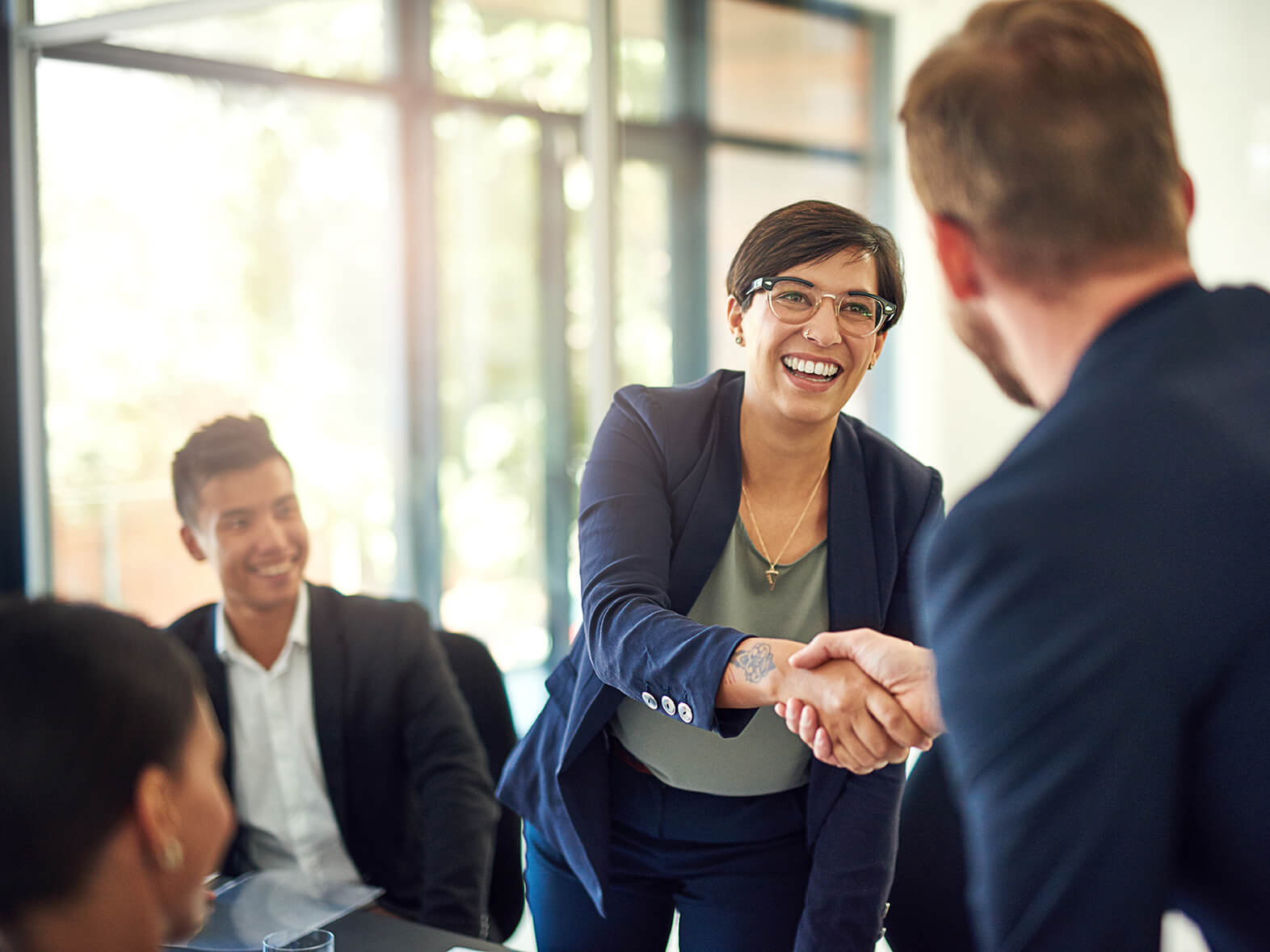 A woman leaning over a desk in a business meeting to shake a colleague's hand 