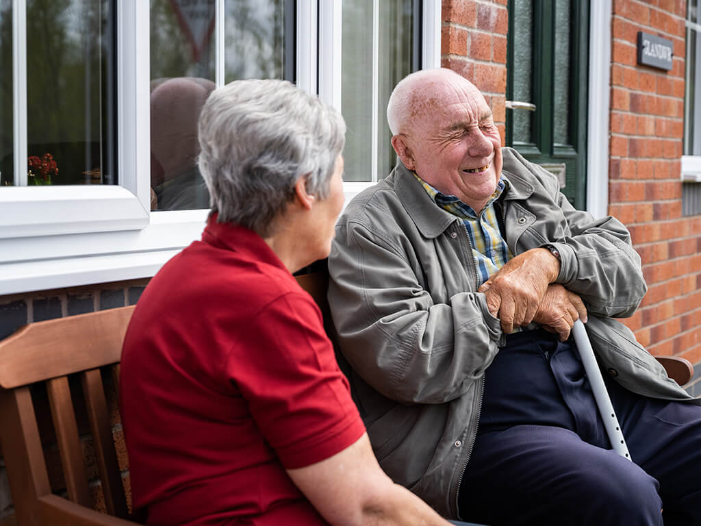 An older man sitting on a bench laughing with a Royal Voluntary Service volunteer
