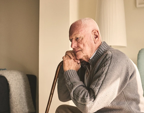 An older man in his home leaning on a walking stick