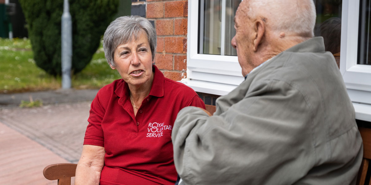 An older Royal Voluntary Service volunteer chatting to a client while sitting on a bench 